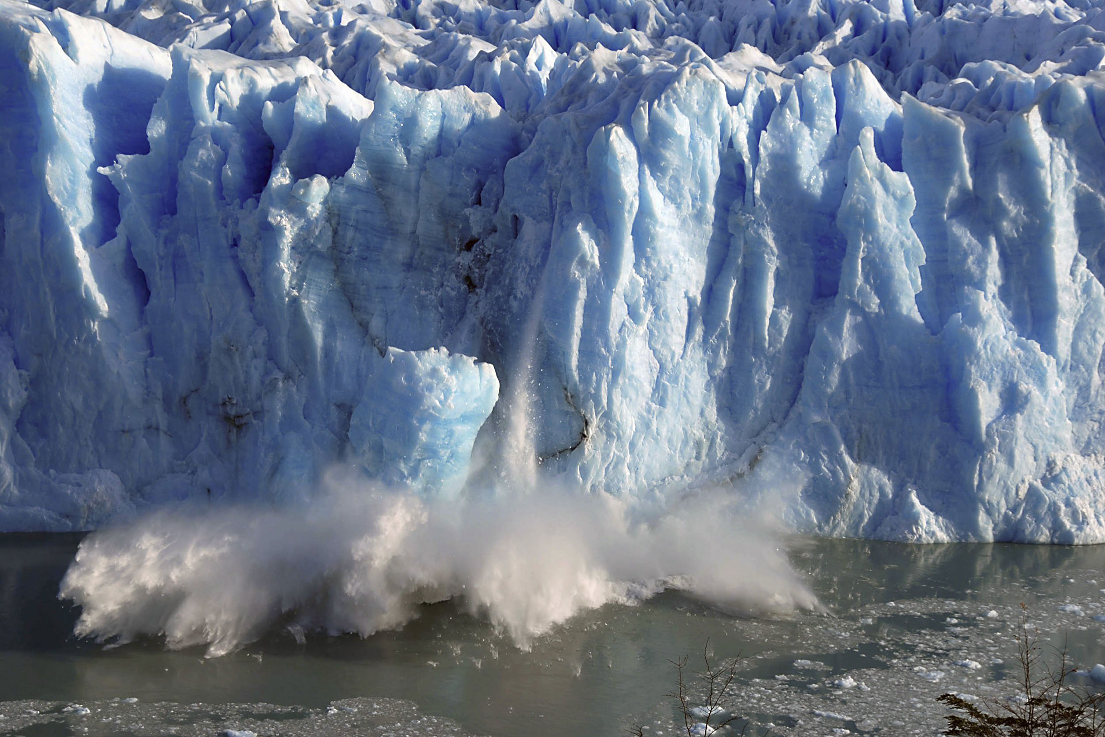 Blue glacier wall with ice breaking off and splashing into cold water below